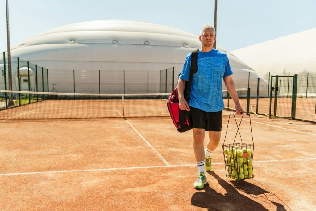 A man on a tennis court carrying a bag and a basket of tennis balls, ready for practice under the b